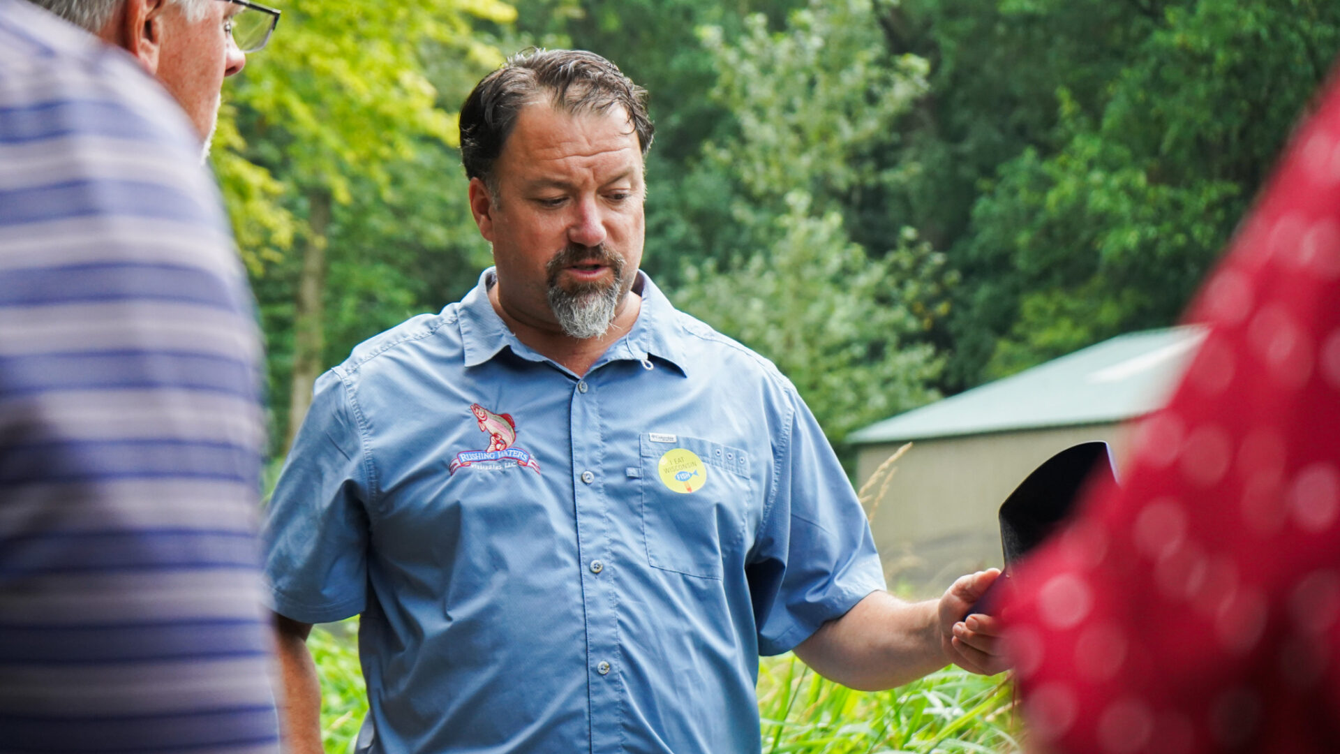 Trout farmer Peter Fritsch wears a blue shirt emblazoned with the logo of his business, Rushing Waters Fisheries, and discusses his business with tour group