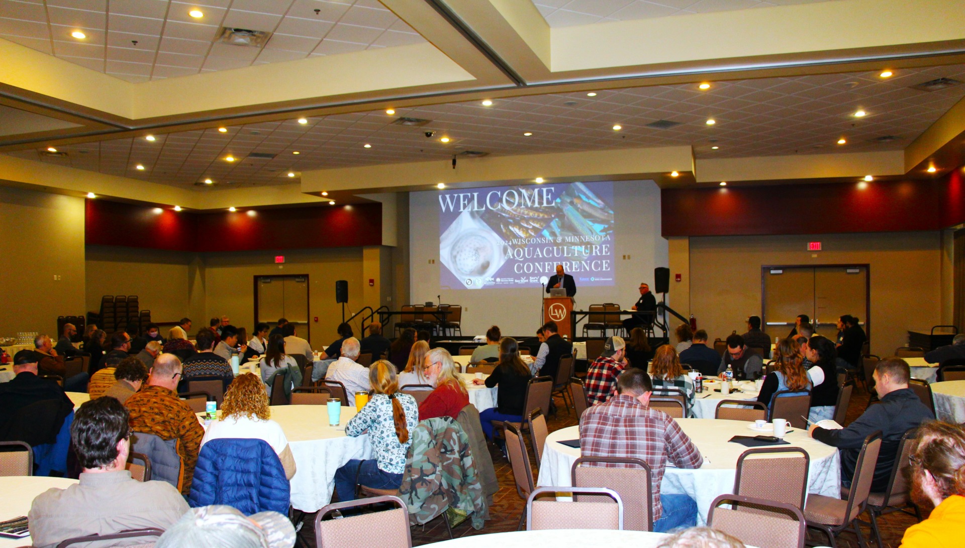 People it at several round tables in a conference room and listen to a speaker at a podium.