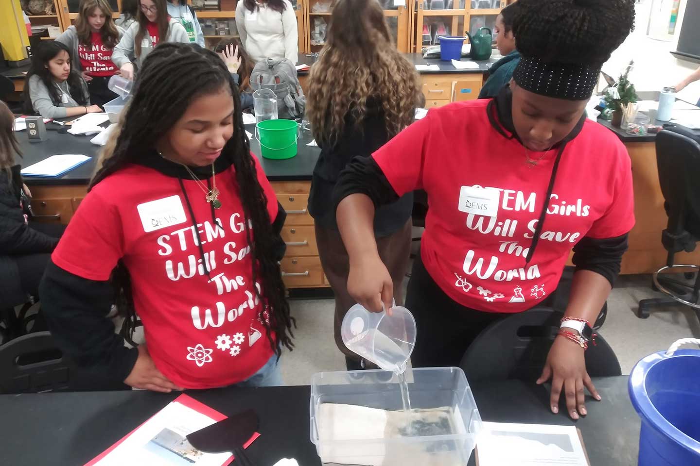 Two students in red t-shirts that say "STEM girls will save the world" pour water into a sand-filled plastic container to prepare for the wave tank activity.