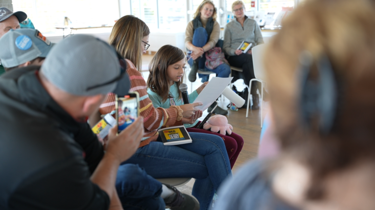 A young girl reads the play while an adult holds a microphone to her. A man is in the foreground filming on his camera phone.