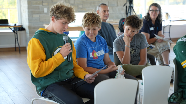 A young man wearing a Packers hoodie is holding a microphone and reading from a piece of paper while seated. Several onlookers watch from his left.