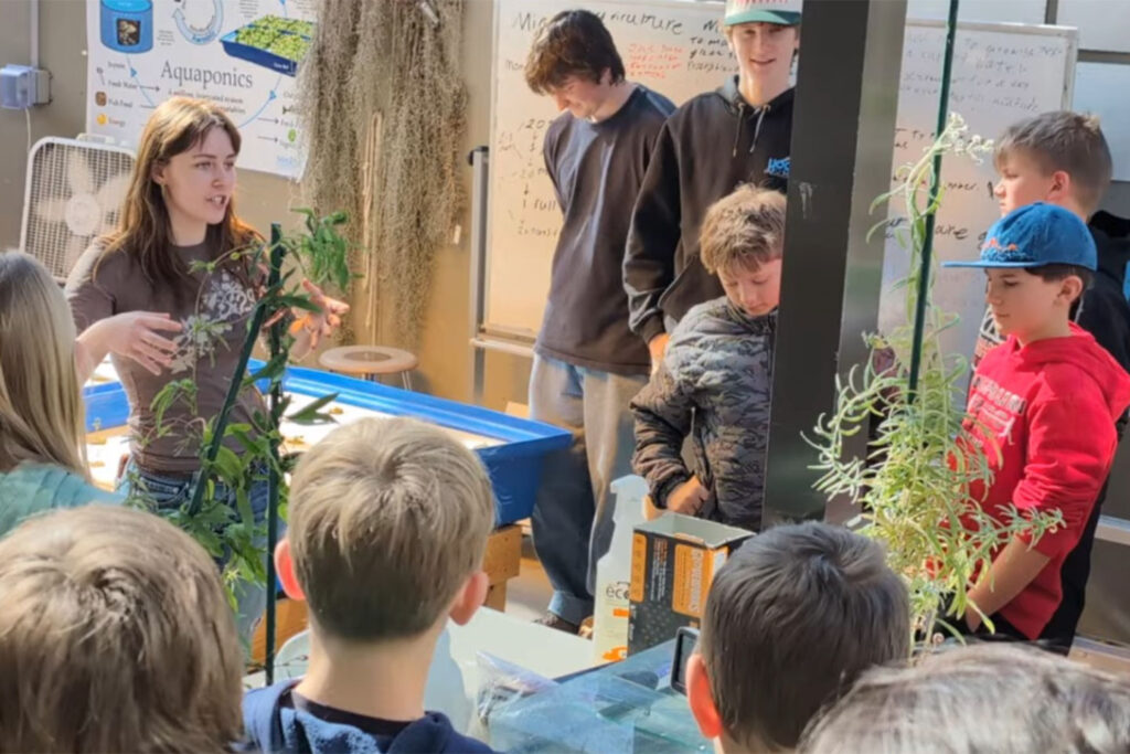 A high school student with long brown hair stands in a science lab and explains aquaponics to a group of sixth graders. Around them, plants crawl up tall stakes.