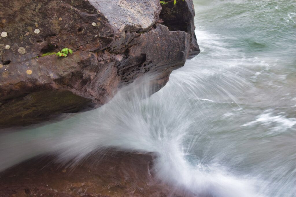 Water from a breaking wave crashes into a rock outcropping overhanging the lake.