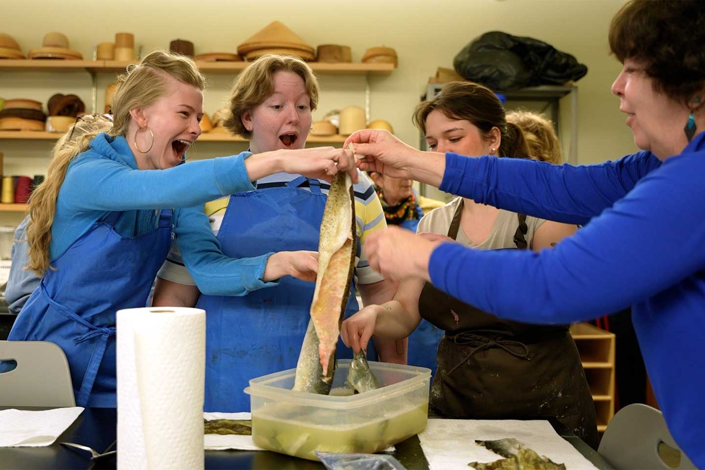 Three surprised and enthusiastic students lift fish skin out of a plastic container with the assistance of an instructor.