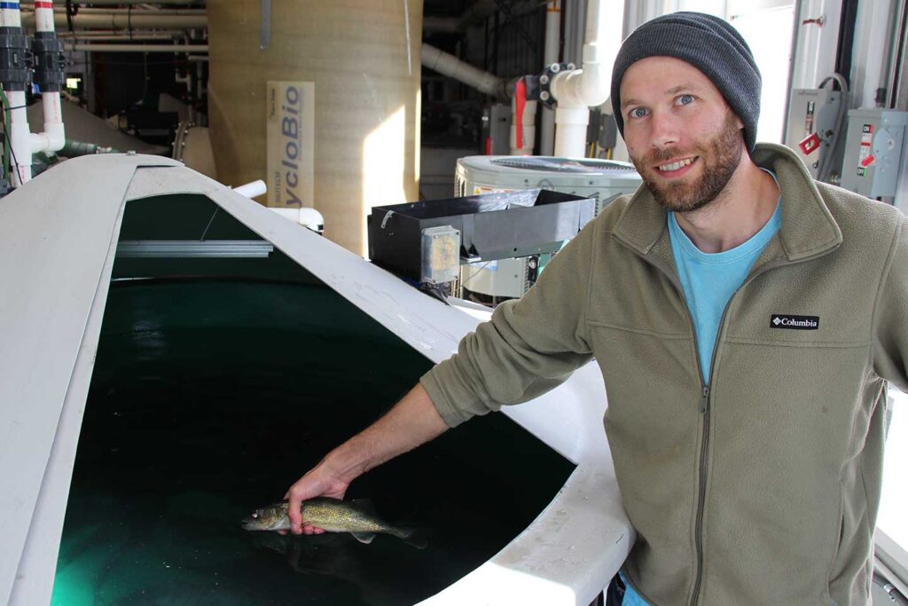A researcher stands beside a large circular fish-rearing tank, holding a walleye. Pipes, filtration equipment, and tanks are visible throughout the indoor aquatic research facility.