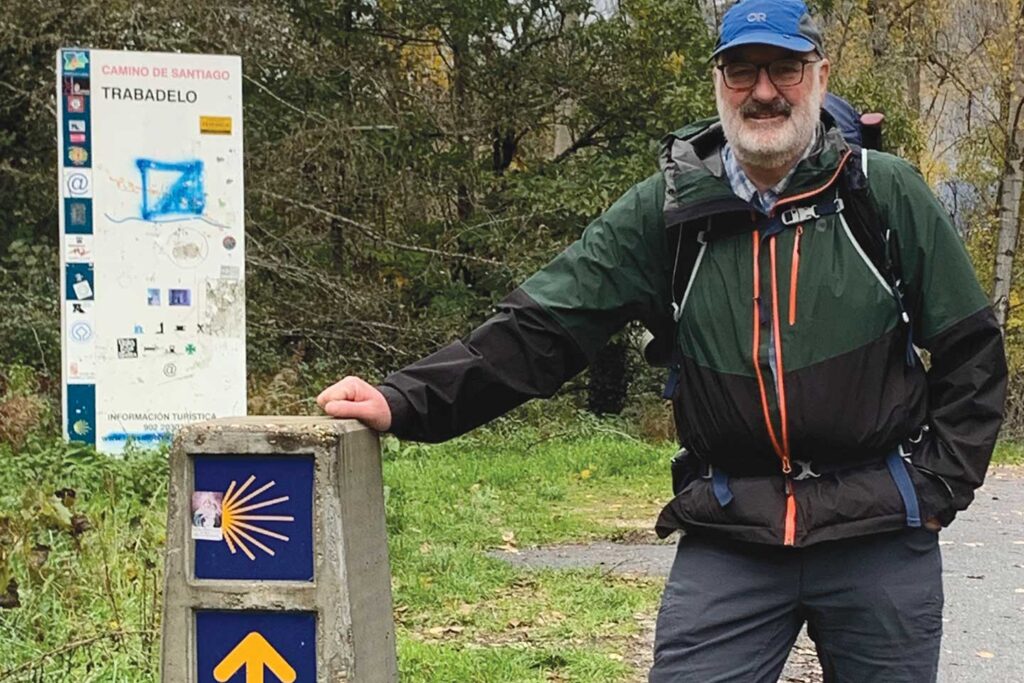 A man wearing hiking clothing and a backpack rests his hand on a trail sign.