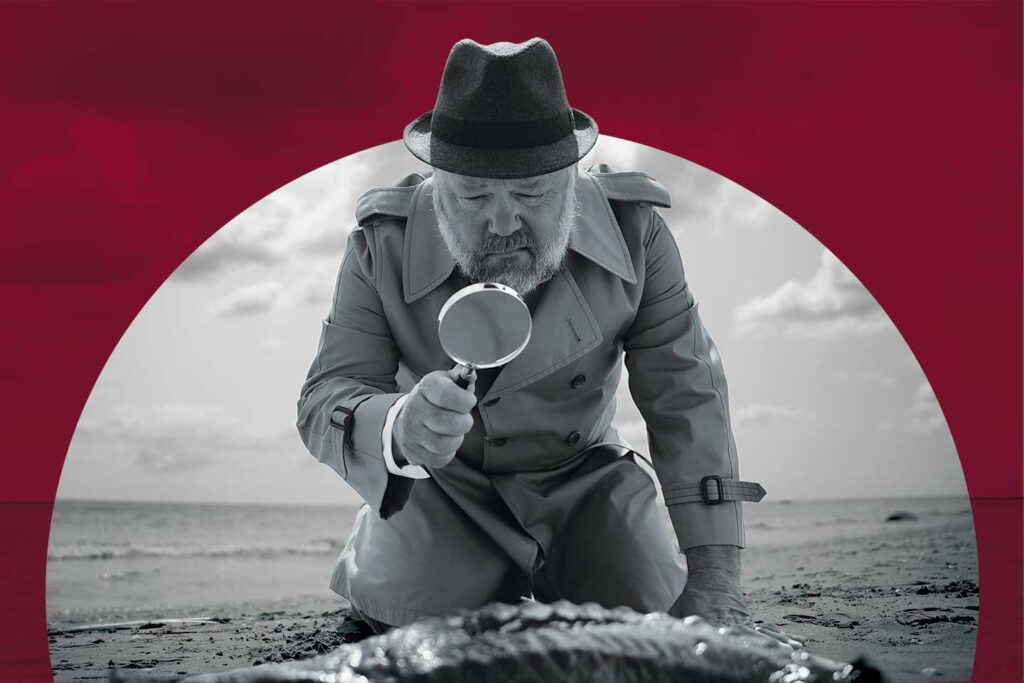Black-and-white image of a man wearing a hat and trenchcoat kneeling on a beach using a magnifying glass to look down at a dead fish.
