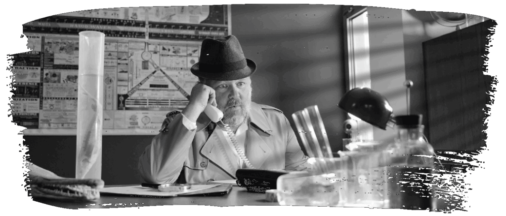 A man sits at a desk surrounded by glass beakers and scientific specimens, holding a phone and wearing a surprised expression.
