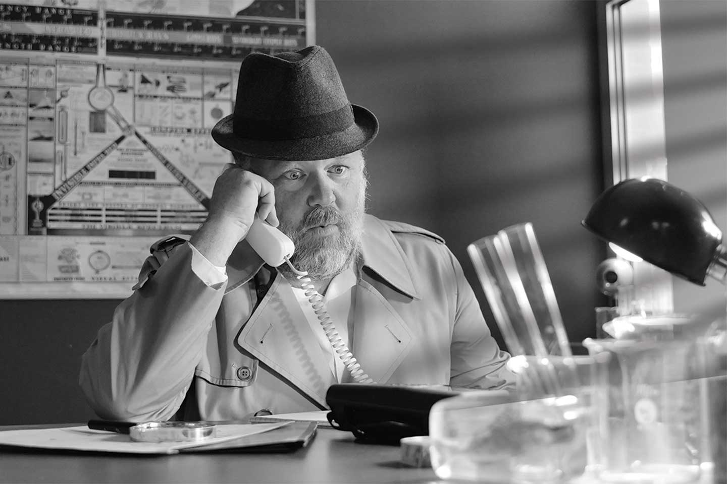 A man sits at a desk surrounded by glass beakers and scientific specimens, holding a phone and wearing a surprised expression.