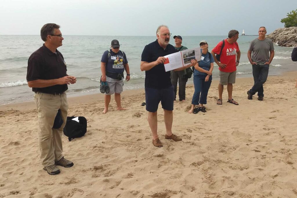 A group of five people standing on a beach with another person in front who is holding a book and talking.