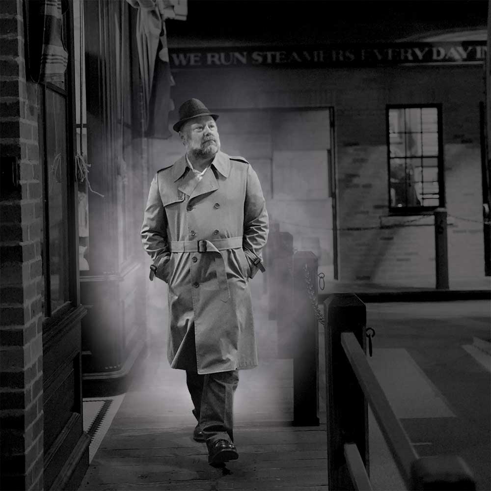 Man wearing hat and trenchcoat strolls pensively in a vaguely foggy alley.