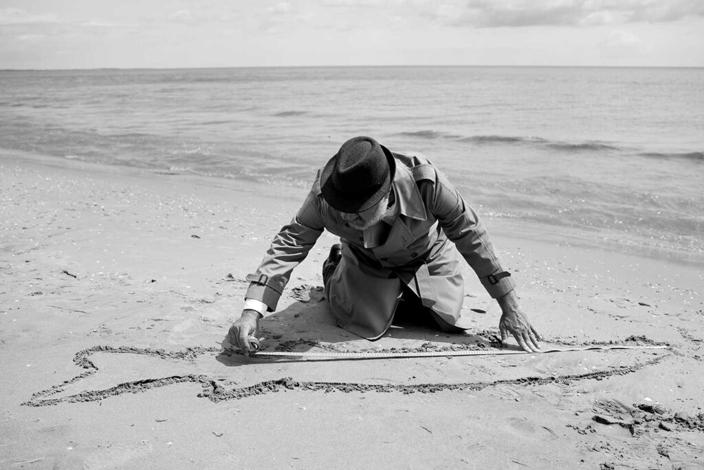 A man wearing a hat and trenchcoat measures an outline of a fish on a beach.