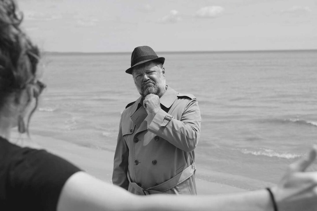 Man wearing hat and trenchcoat stares into the camera with a quizzical expression with another person facing him partially visible.