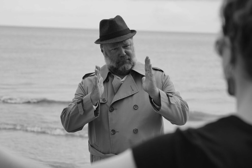 Man wearing hat and trenchcoat holds hands in front of chest indicating size of fish with another person facing him partially visible.