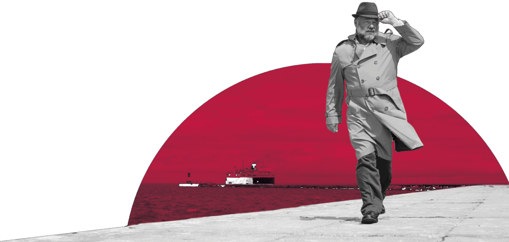 A man wearing a hat and trenchcoat tips his hat as he walks down a pier with a ship behind him.